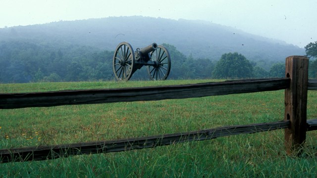 A cannon in a filed behind a wooden fence.