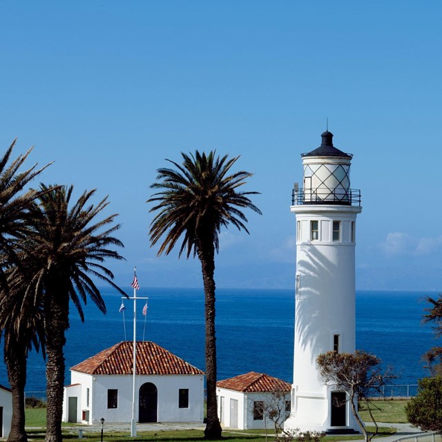 A lighthouse and keepers quarters with red tile roofs with blue water and sky behind. 