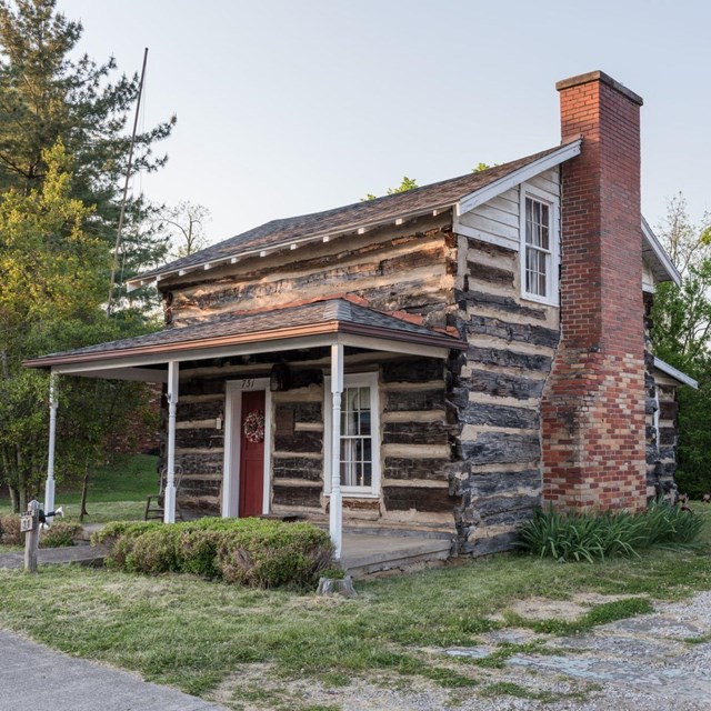 An oblique view of a log structure with a front porch and a red brick chimney