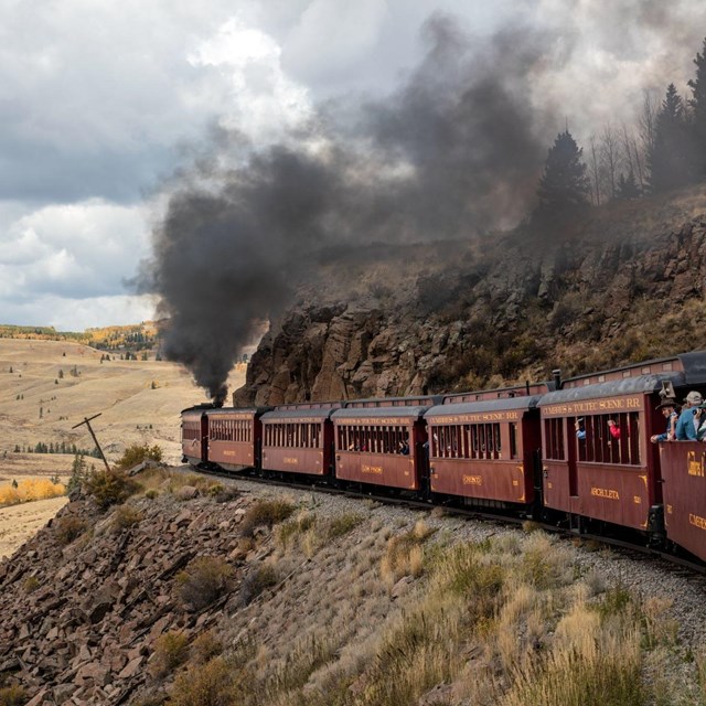 A red train travels along tracks built into a shelf on a hillside with smoke streaming from a stack