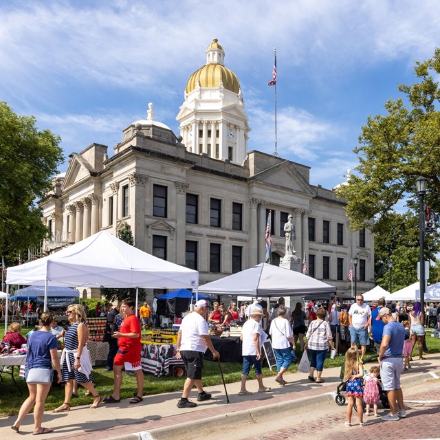 People attend a street fair held in front of a historic, stone, three-story building.