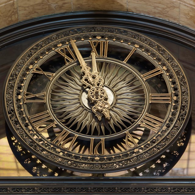 The face of a clock in a stone arch has brass hands pointing to 2 minutes to twelve. 