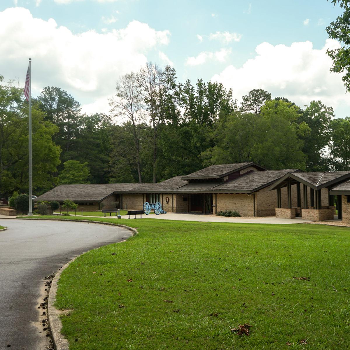 The image displays a brick building, with a concrete walkway and replica cannon in front of it.