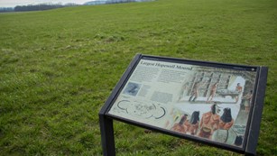 An interpretive panel exhibit displaying info and pictures in front of a grass-covered mound