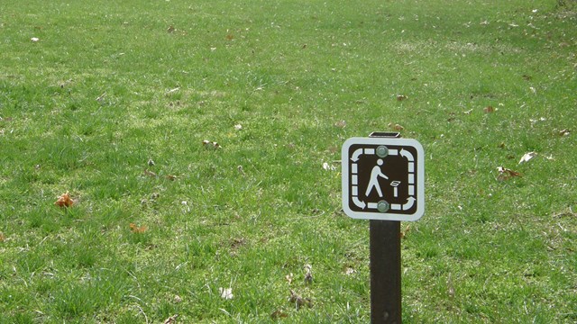 A small brown trail sign in front of a wide area of grass on a trail