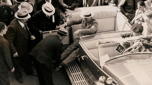 An eager crowd watches as Secret Service agents straighten and lock FDR’s leg braces in preparation.