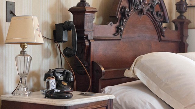 A bed with nightstand and wall-mounted telephone.