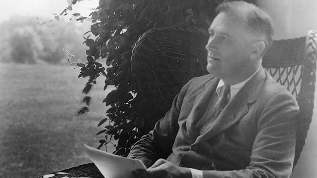 A man seated outdoors on a veranda with river in the distance.