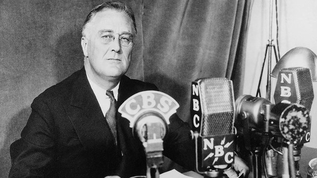 A man in a suit at a desk with many microphones.