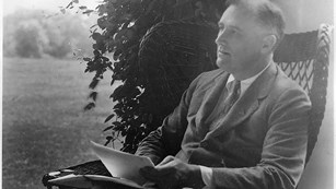 A man in a suit with papers in his hand, seated on a porch.