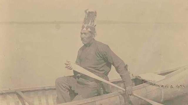 An elderly man with feathered head dress navigating open waters in a conoe.