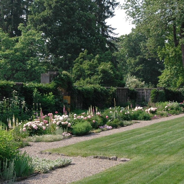 An open lawn surrounded by garden beds of blooming flowers in front of a vine covered stone wall.