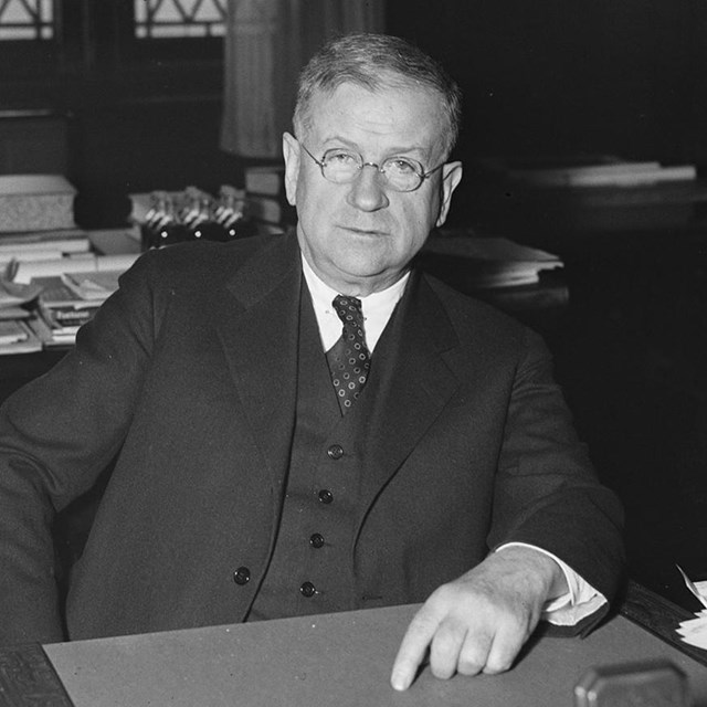 A man in a dark suit seated behind a desk with papers.