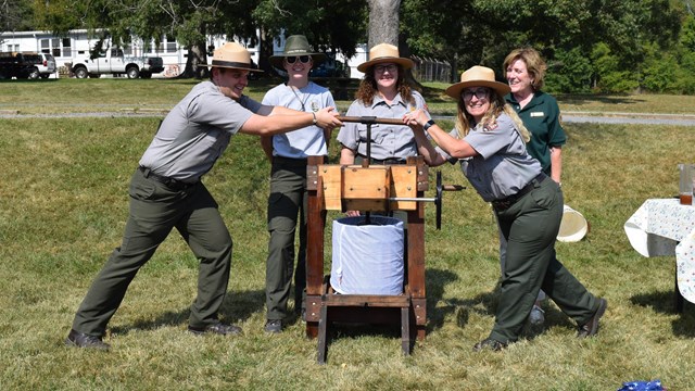 A group of rangers and a volunteer pose by a large wooden apple cider press. 