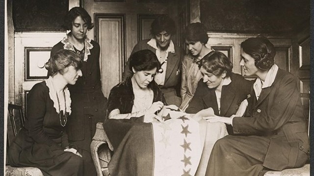 Women watching another woman sew a star on a National Women's Party flag.