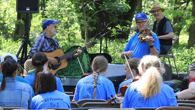 Two men stand in front of an outdoor class. One plays the fiddle. The other plays guitar.