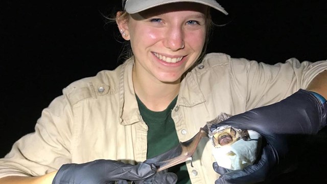 Girl holds bat. She is smiling and wearing a ball cap and black rubber gloves.