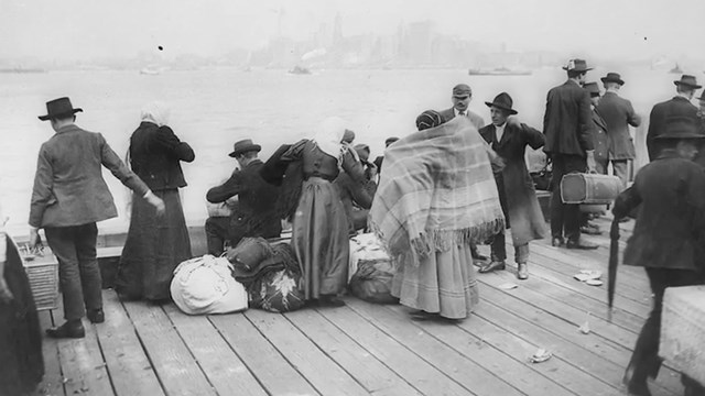 Men and Women wait on a dock, New York City looms in the distance across the bay.