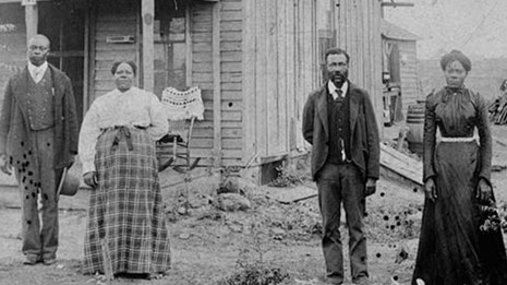 Two black men and two black women stand in front of a frame house. Photo is black and white.