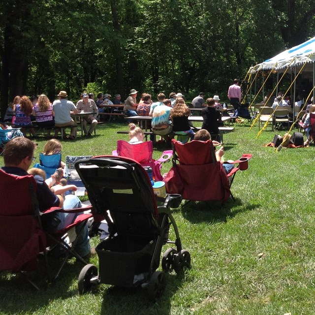 A crowd of people sit in lawn chairs and at picnic tables. There is a tent with people in it.
