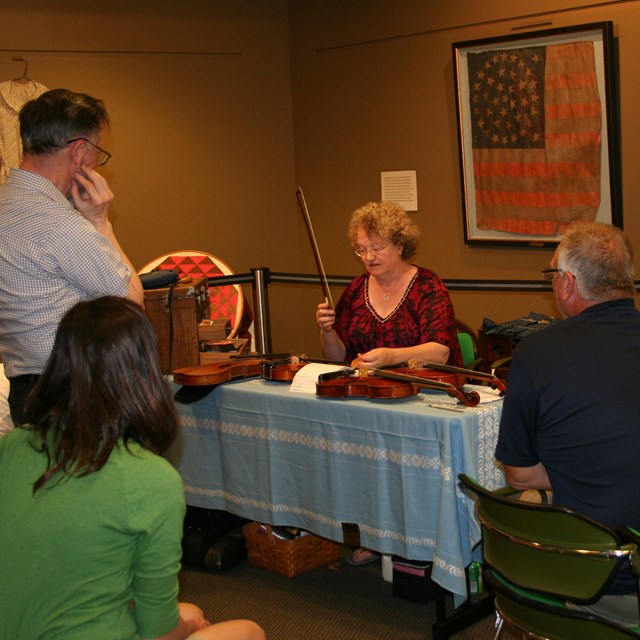 A woman sitting at a table holds a fiddle and bow while others look on.