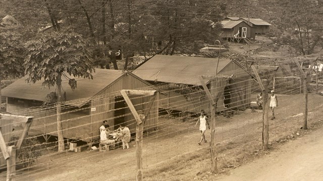 photo of people sitting at a table outside wooden buildings behind a tall barbed wire fence