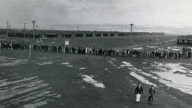 photo of a long line of people standing in front of many wooden buildings in a flat, barren land