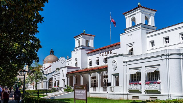 An active scene in front of the Fordyce Bathhouse; people sitting ,walking, and looking joyous.