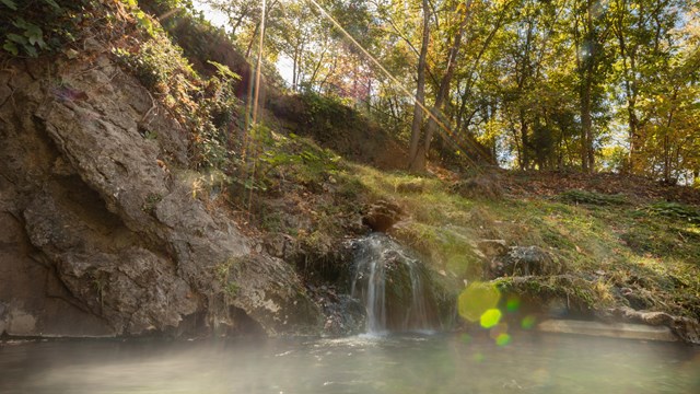 Water cascades over a hillside and into a thermal pool with the sun shining through trees.