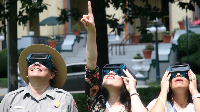 A woman in a ranger uniform and two visitors look up at the sky in awe while wearing eclipse glasses