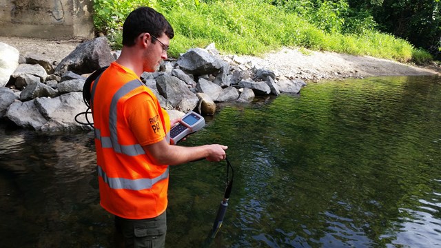 A man dressed in bright orange holds monitoring equipment in a stream.