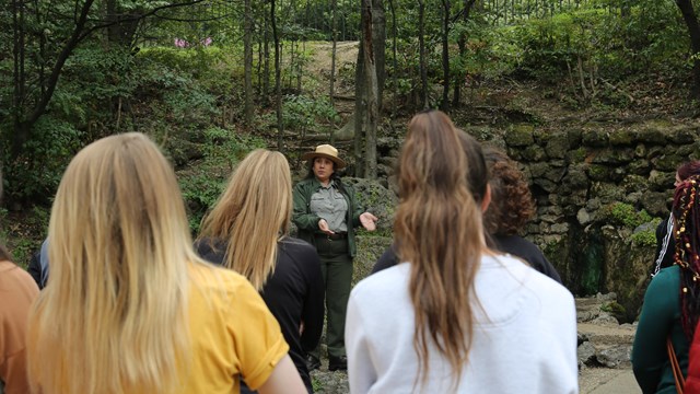 A woman in a gray shirt, green jacket, green pants, and straw hat stands in front of a group.