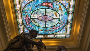 Ornate stained glass in the Fordyce Bathhouse with a statue of Hernando De Soto in the foreground.