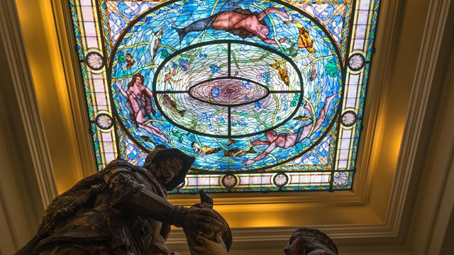 Ornate stained glass in the Fordyce Bathhouse with a statue of Hernando De Soto in the foreground.