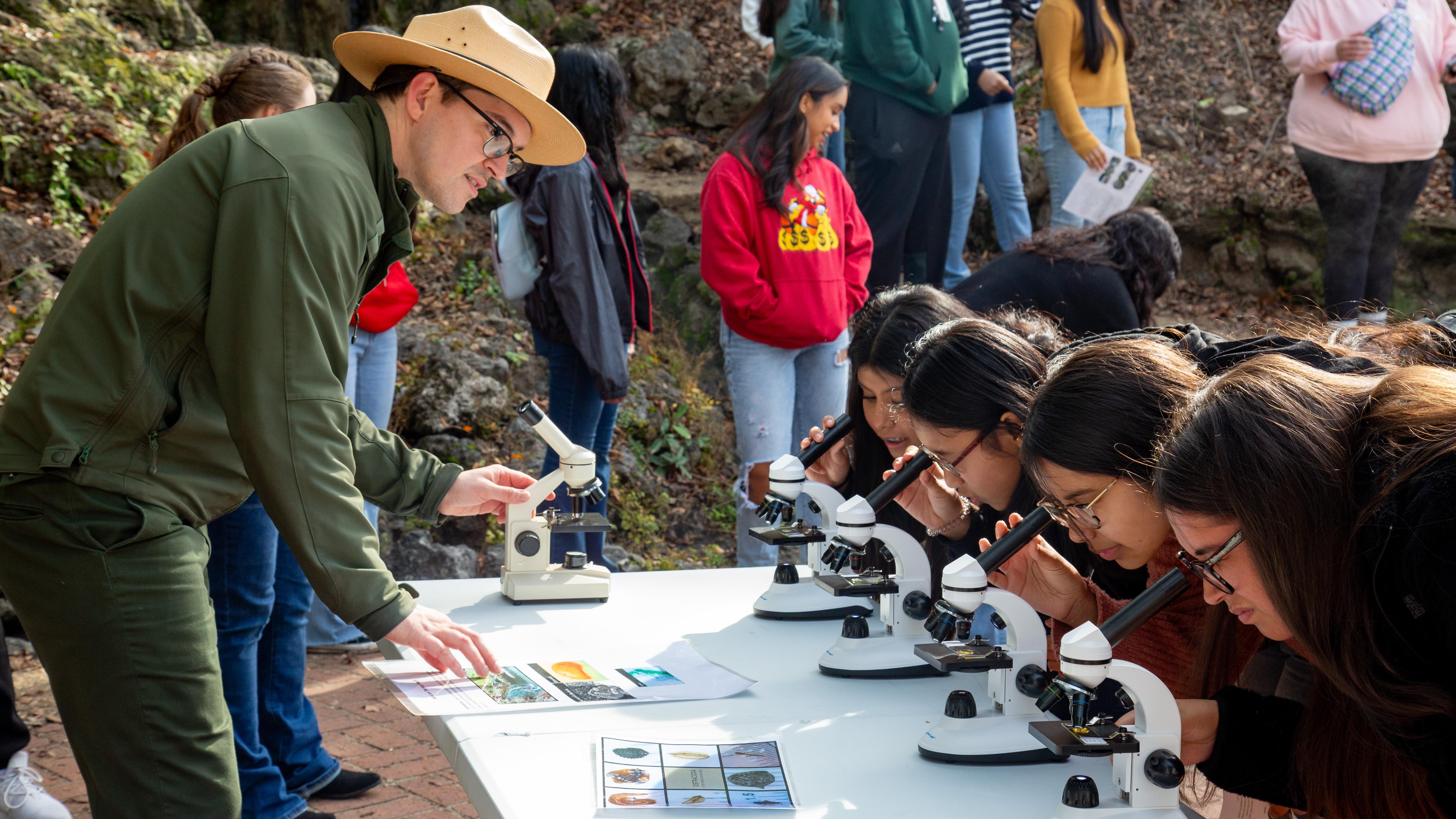 A tall ranger stands at a table with kids looking through microscopes.