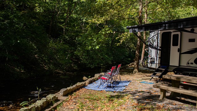Two camp chairs next to an RV by the creek.