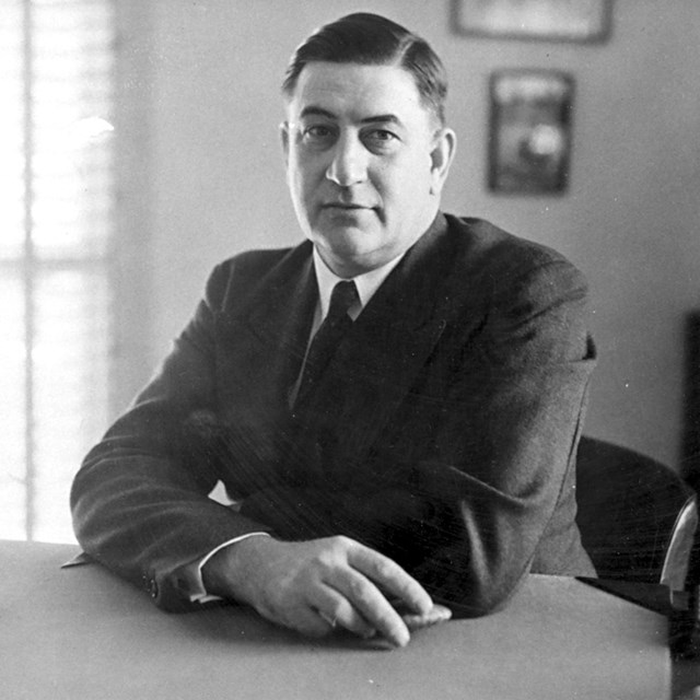 A gentleman sits in an office at a desk, arms crossed looking straight into the camera.