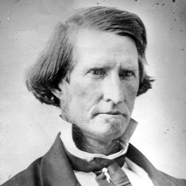 A man with parted long hair to his ears with a stern face looks towards the camera.