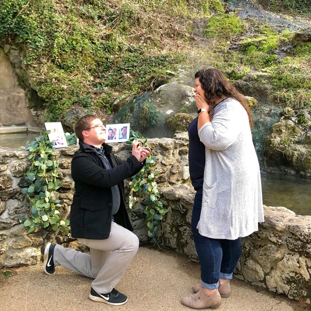 A man is on his knee proposing to a woman in front of a display spring.