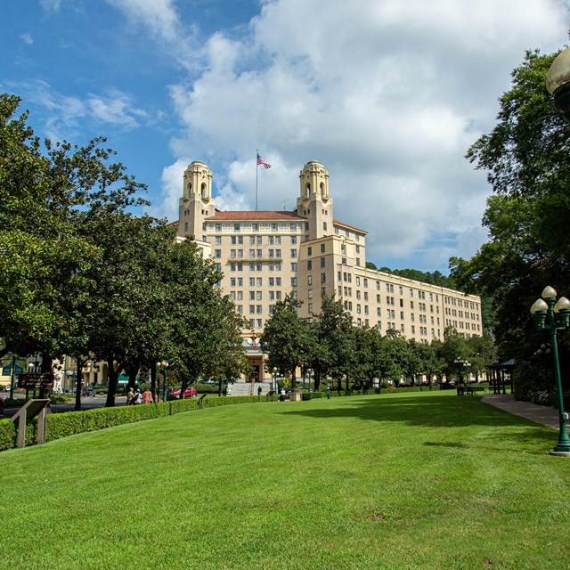 A tall and wide tan building sits in front of a bright blue sky and behind a lush green lawn.