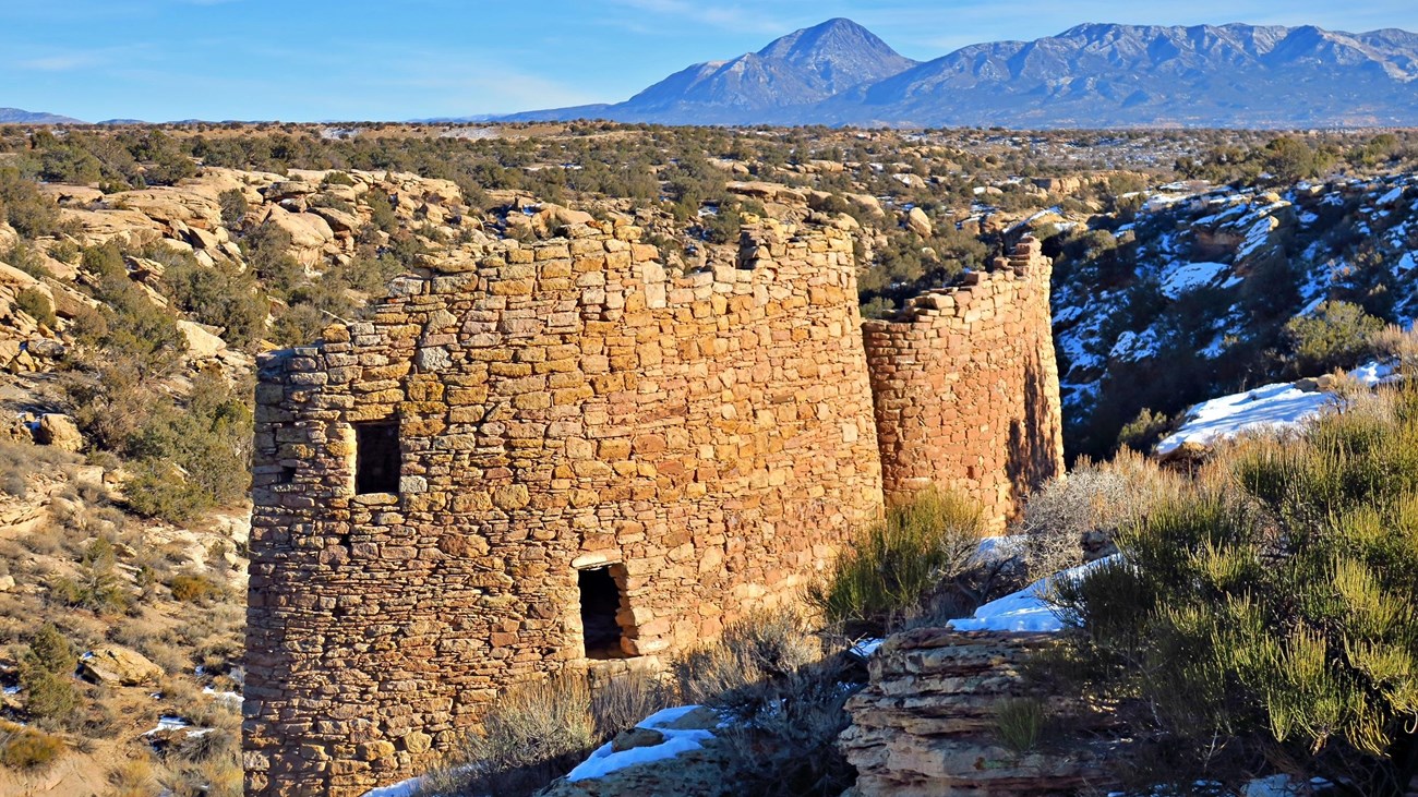 An old stone structure with two windows stands near patchy snow in a canyon with mountains beyond it