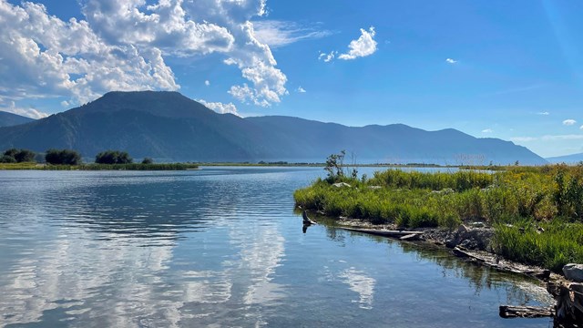 A lake reflecting coulds with a green mountain in the background