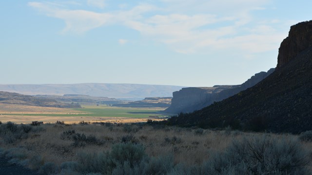 dark canyon walls give way to a sunny distant landscpe of a flooded coulee.