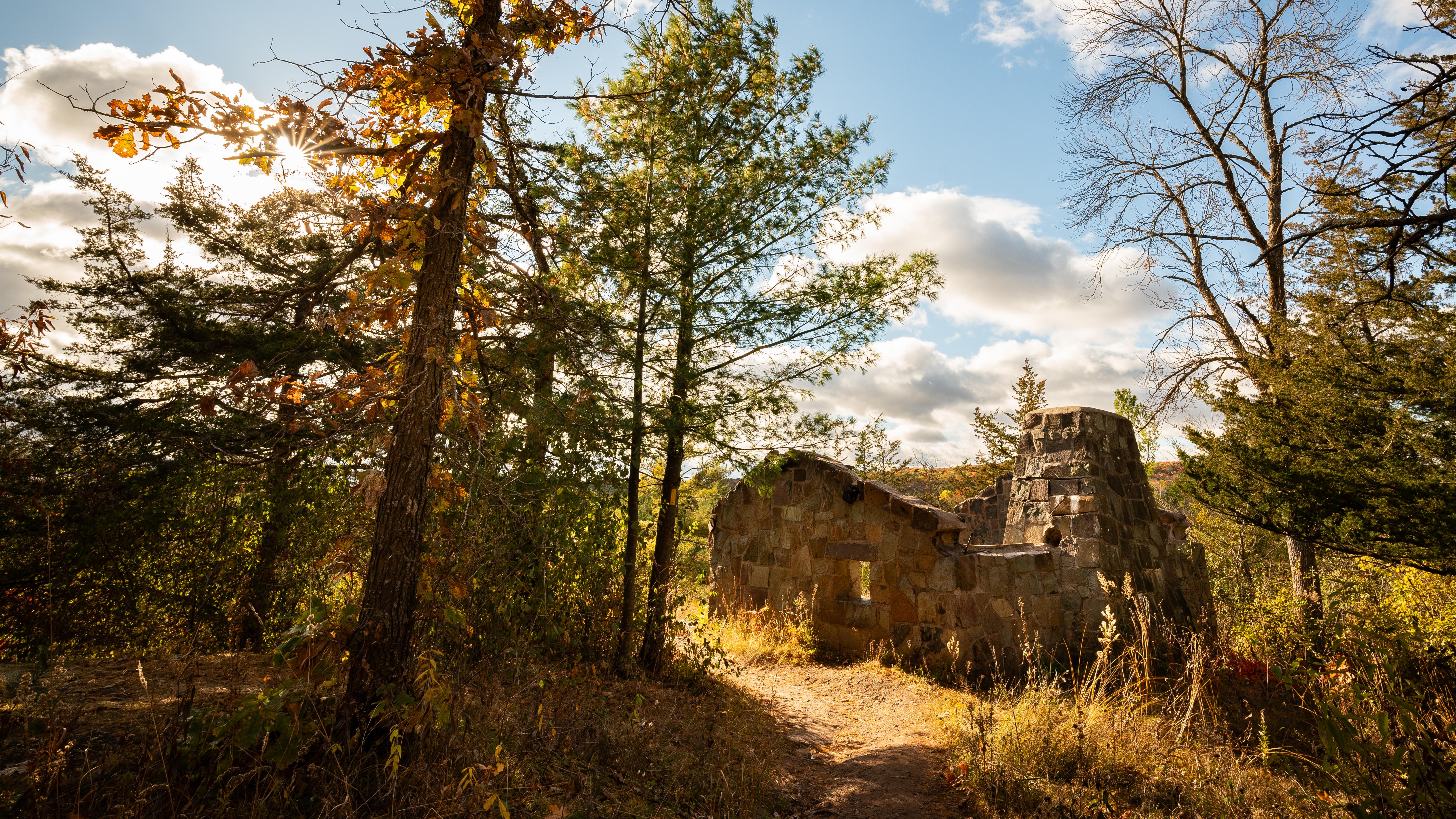 Photograph of trail near stone building ruins surrounded by trees under a partly cloudy sky. 