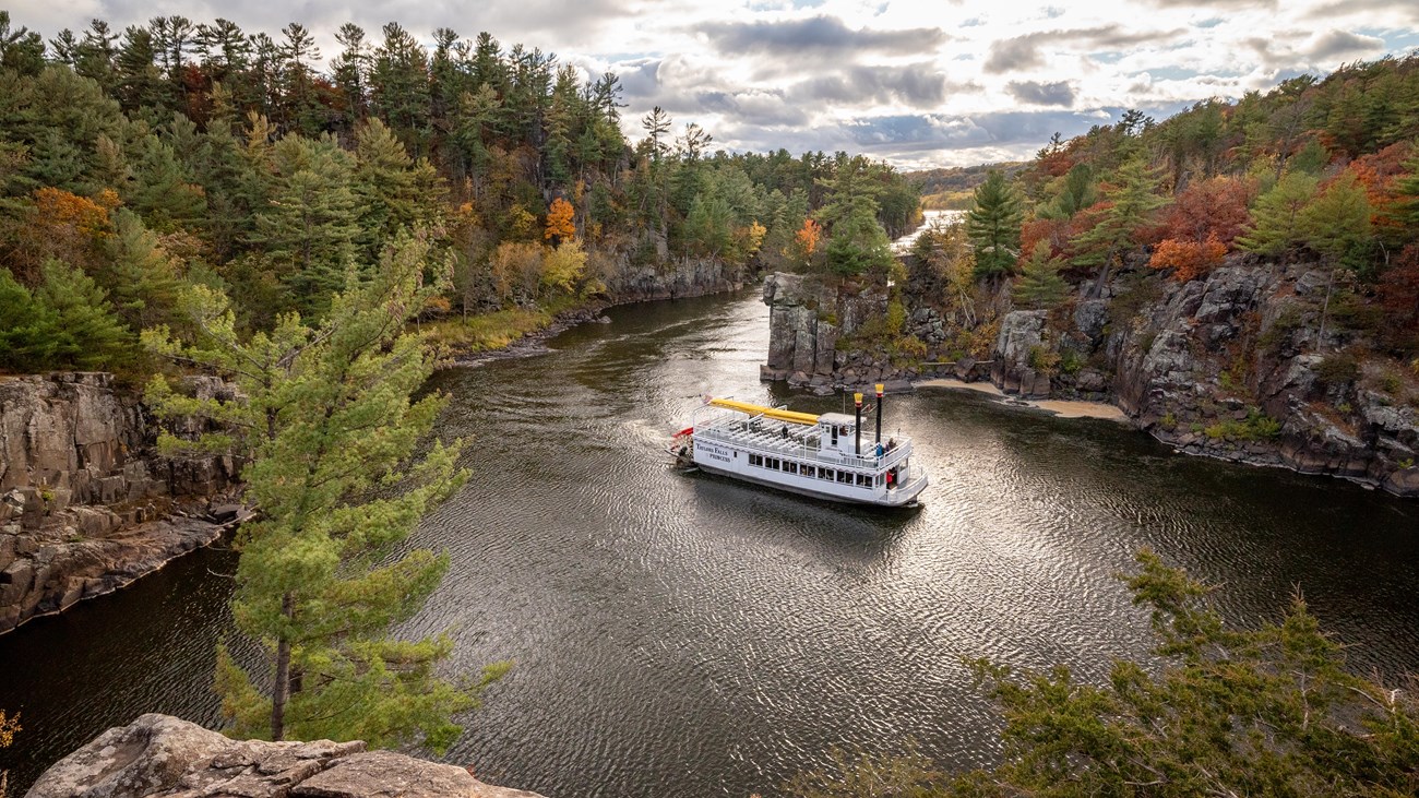 Photograph large paddleboat rounding a bend in a river with steep rock walls and trees. 