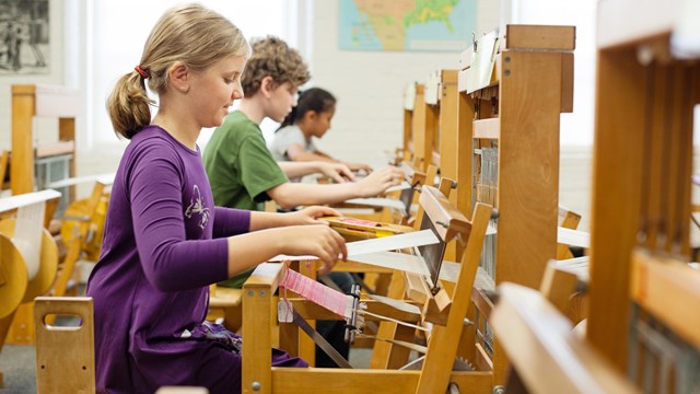 Three children sit at wooden looms, weaving cloth