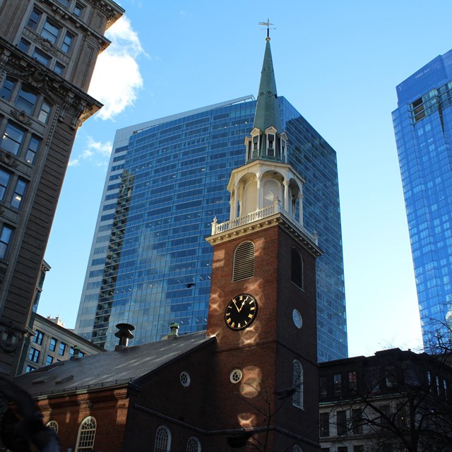 tower of dark red brick building, with a clock in the middle, surrounded by modern buildings 