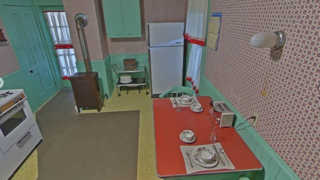 Inside a kitchen with patterned wallpaper, a red topped table with a toaster and two place settings.