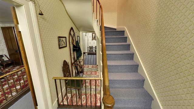 A narrow hallway separates a carpeted staircase with a wood banister from a small living room.