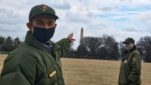 Two rangers in face masks, one pointing to the Washington Monument